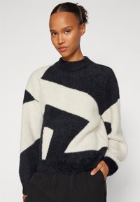 Pulover supradimensionat dintr-un tricot pufos, cu un design geometric în alb și negru, guler înalt și accente de model texturat.
