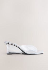 Prozirna natikača s potpeticom, dizajnirana s zakrivljenim oblikom, izrađena od prozirnog PVC materijala i sjajnih metalik detalja, s kvadratnim vrhom i potpeticom visine pola inča.