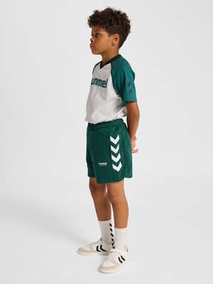 Chłopiec o kręconych włosach stojący bokiem, ubrany w szaro-zielony strój sportowy, białe skarpetki i białe trampki z czarnymi logo w kształcie szewrona.