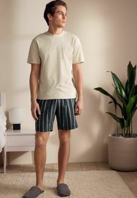 Hellbeige Herren-T-Shirt mit Brusttasche, kombiniert mit dunkel gestreiften Shorts. Graue Hausschuhe. Inneneinstellung mit einer Pflanze und einer Lampe.