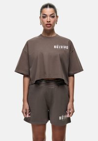 T-shirt cropped marrone e pantaloni coordinati realizzati in tessuto morbido. Presenta un logo stampato in bianco sul lato sinistro del petto e sull'orlo dei pantaloni.