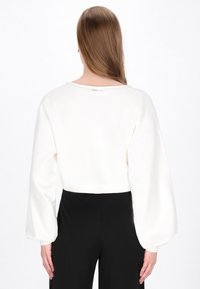 Pull féminin blanc court avec des manches longues et bouffantes, un col rond, en tissu texturé et avec une petite ouverture au dos. Assorti avec un pantalon noir.