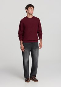 Maglione lavorato a maglia bordeaux con polsini e orlo a coste, abbinato a jeans dritti grigio scuro e scarpe marroni. Silhouette semplice e tessuto testurizzato.