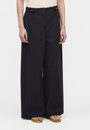 Person iført vidde sorte bukser med snor i taljen og lysegrønne flip-flops, stående mod en ensfarvet baggrund.