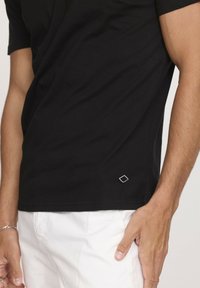T-shirt en coton noir avec un col rond, des manches courtes et un petit logo en forme de diamant en bas à gauche de l'ourlet. Associé à un pantalon blanc.