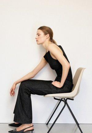 Joven mujer con top negro sin mangas y pantalones sentada de lado en una silla beige, inclinándose hacia adelante con una mano apoyada en la rodilla.