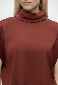 Femme aux cheveux blonds et lisses portant un haut sans manches à col montant, couleur rouille, vue des épaules au menton sur un fond uni.