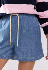 Shorts in denim con una superficie leggera, con elastico in vita e cordoncino crema con estremità dorate. L'orlo presenta dettagli di cucitura chiara.