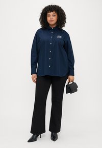 Marineblå knapp-up skjorte med et merket logo, kombinert med svarte vide bukser og en liten svart håndveske. Høye svarte sko fullfører antrekket.