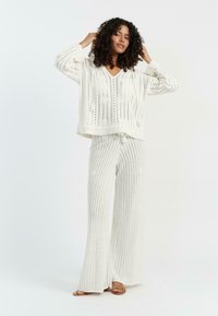 Maglione bianco a maglia con scollo a V e dettagli con frange abbinato a pantaloni a gamba larga a righe. Entrambi presentano un design testurizzato e a trama aperta.