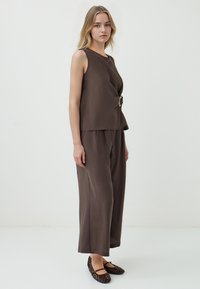 Bruine mouwloze top met een gedraaide design en zilveren hardware-detail, gecombineerd met bijpassende wijde broek en zwarte gevlochten flats.