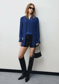 Blusa di satin blu con scollatura a V, abbinata a pantaloni corti neri. La modella indossa stivali neri fino al ginocchio e tiene in mano una piccola borsa nera.