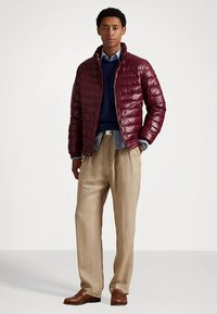 Casaco acolchoado bordeaux com costura horizontal, camisola azul-marinho, camisa azul com colarinho, calças de linho bege e sapatos de pele castanhos.