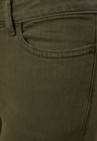 Tela de mezclilla verde oliva con una textura suave, que presenta un bolsillo frontal y un botón de metal. Las costuras son visibles a lo largo de las costuras.
