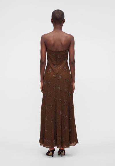 Femme aux cheveux courts portant une robe longue marron bustier avec broderie florale et sandales noires à talons hauts, debout de dos sur un fond uni.