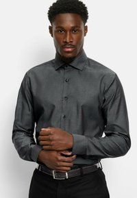 Donkergrijze blouse met een subtiele textuur, met een kraag, lange mouwen en zwarte knopen. Gedragen met een zwarte riem.