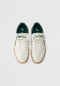 Weiße Sportschuhe mit grünen Akzenten, Obermaterial aus strukturiertem Leder und Mesh, goldenes "Puma"-Logo, verstellbarer Riemen und Sohle aus Gummi.