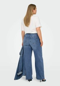 Top ligero blanco con encaje en las mangas, combinado con jeans azul de pierna ancha de talle alto. La modelo sostiene una chaqueta de denim.