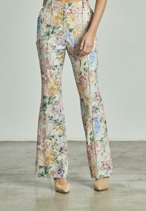 Vrouw in hoge taille, bloemenprint uitlopende broek en beige puntige hakken staat op een gepolijste betonvloer.