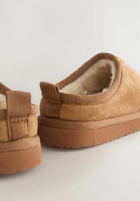 Bruna mockaskor utan snörning med en mjuk, fluffig foder, texturerade detaljer och en gummiyttersula med ett vågigt mönster.