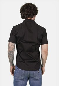 Camicia nera a maniche corte con colletto piatto e spallina, realizzata in tessuto morbido, indossata con jeans blu, mostrando un tatuaggio dettagliato sul braccio.