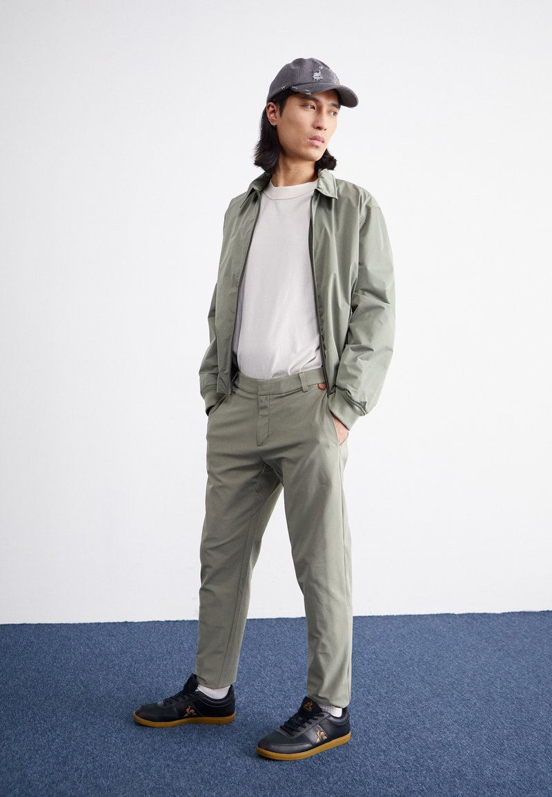 Camiseta de color gris claro debajo de una chaqueta con cremallera en verde salvia, combinada con pantalones verde claro y zapatillas negras con suelas de goma sobre una alfombra azul.