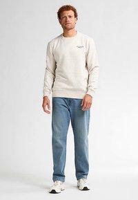 Hellbeiges Sweatshirt mit Sprenkeln, Rundhalsausschnitt und eng anliegenden Bündchen. Kombiniert mit hellblauen Jeans und weißen Sneakern. Minimalistische Designelemente.