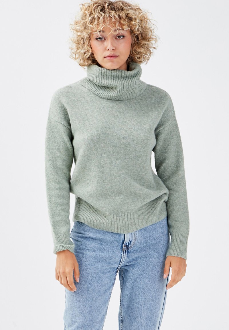 Suéter de cuello alto en verde claro, hecho de una tela suave y texturizada. Con mangas largas y un corte holgado, combinado con jeans azules.