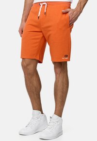 Orange svedshorts med elastisk talje og snørelukning. Har sidetekster og et logo på venstre lår. Bæres med hvide høje sneakers.