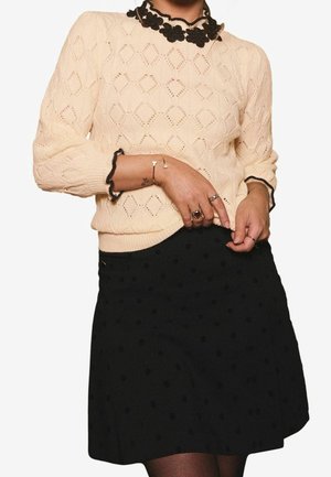 Kremowy sweter dzianinowy z diamentowymi wzorami i czarnym kołnierzem z kwiatowej koronki, zestawiony z czarną spódnicą w kropki oraz pierścionkami na palcach.