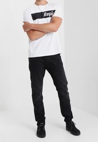 Vit t-shirt med svart horisontell band och texten "Repl", i kombination med svarta jeans och svarta sneakers. Enkel design, rak passform.