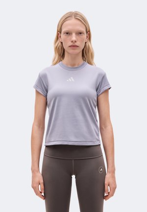 Жена стои с лице напред, облечена в светлолилава тениска Adidas и тъмен клин, на фона на изчистен светъл фон.