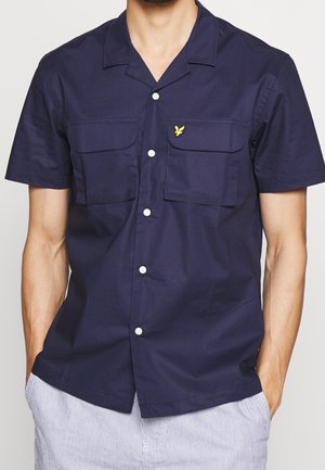 Hombre con camisa azul marino de manga corta y botones, con dos bolsillos en el pecho, uno de los cuales tiene un pequeño logo de pájaro amarillo.