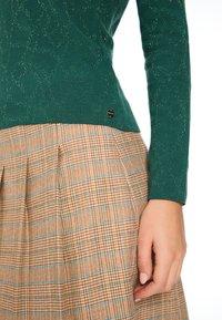 Grüner Strickpullover mit strukturiertem Muster und metallischen Fäden, kombiniert mit einem braunen karierten Rock mit blauen Akzenten und Falten.