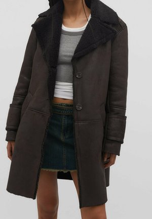 Femme portant un manteau en peau lainée marron foncé sur un crop top gris côtelé et une mini-jupe en denim à ourlet effiloché, debout devant un fond uni.