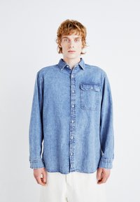 Cămașă din denim albastru deschis cu guler cu nasturi, mâneci lungi, un singur buzunar pe piept și o croială lejeră. Prezintă o textură decolorată.