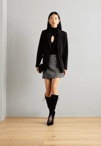 Blazer negru, pulover negru strâmt cu decupaj, fustă mini din tweed și cizme negre cu șireturi până la genunchi. Texturi netede și croială ajustată.