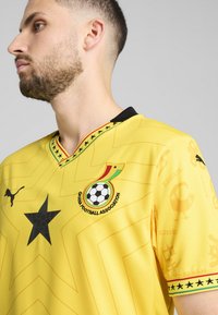 Žltý futbalový dres s výstrihom do V, čiernymi akcentmi, veľkou čiernou hviezdou a logom Ghanskej futbalovej asociácie, s jemnými geometrickými vzormi.