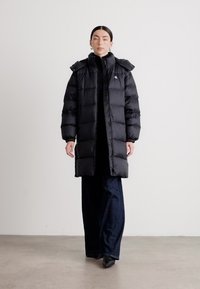 Lang svart dunjakke med hette, med et kviltet design, frontglidelås og side lommer. Båret over en svart topp og mørk denimbukser.
