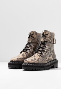 Bottes cheville à motif peau de serpent en beige et noir, avec une semelle épaisse noire, un laçage à l'avant et un détail de boucle sur la sangle supérieure.