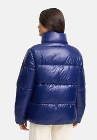 Granatowa kurtka puchowa z wysokim kołnierzem, błyszczącą fakturą, poziomymi przeszyciami i naszywką z logo na rękawie.