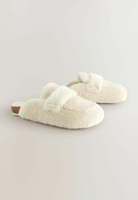 Pantofole color crema realizzate in materiale morbido e peloso con una superficie testurizzata, caratterizzate da una fascia sulla parte superiore e una suola piatta.