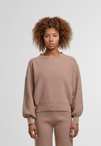 Pull en tricot beige avec de longues manches amples et des poignets côtelés. Tissu texturé et col rond. Associé à un pantalon en tricot assorti.