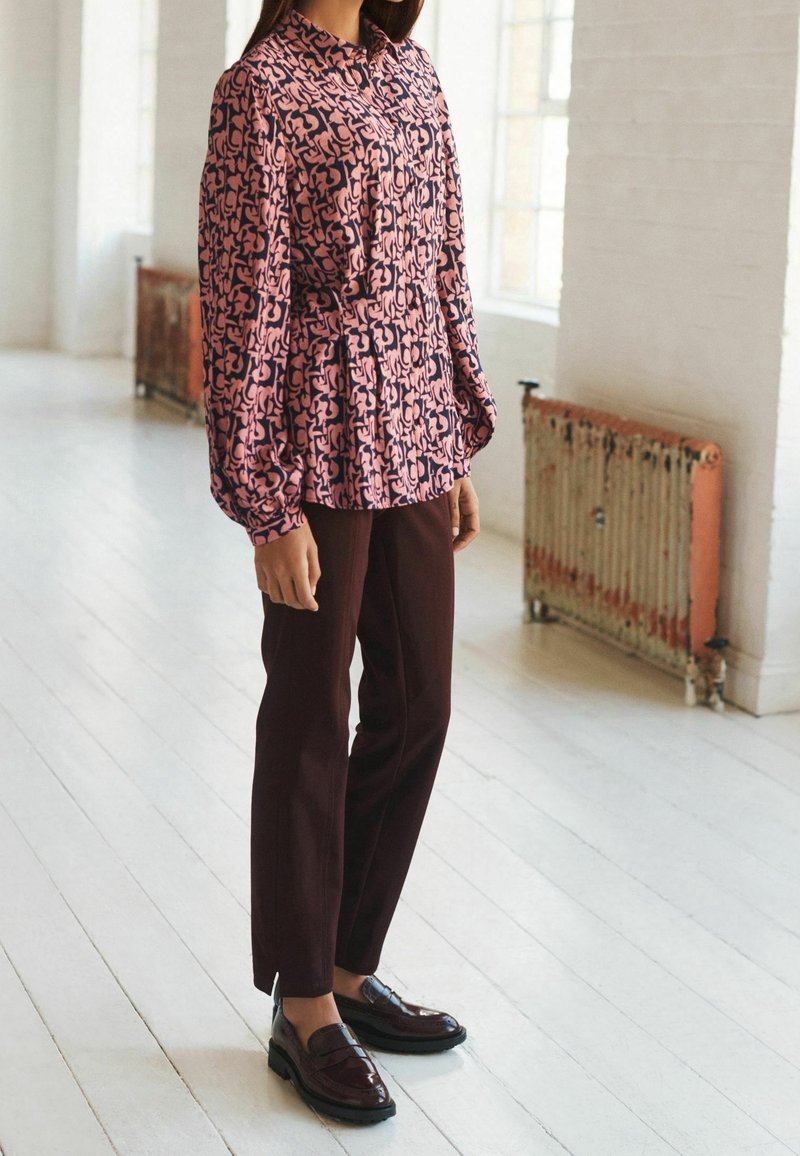 Blouse à motifs rose et marine avec un col et une texture plissée, associée à un pantalon slim bordeaux et des mocassins foncés brillants.