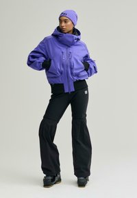 Frau trägt eine lila Mütze und Jacke, schwarze Skihose und Skischuhe, steht mit den Händen in den Jackentaschen vor neutralem Hintergrund.