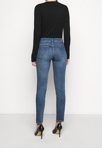 Vaqueros ajustados de color azul con bolsillos traseros y detalles de costura sutil, combinados con una blusa negra de manga larga y tacones altos negros.