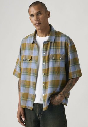 Hombre con camisa de cuadros de manga corta en color mostaza y azul claro sobre camiseta blanca, de pie con una mano en el bolsillo del pantalón vaquero.