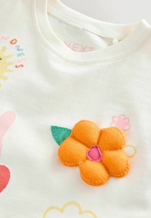 Bijela pamučna majica s aplikacijom narančastog cvijeta s ružičastim središtem i zelenim listom, okružena šarenim printanim uzorcima.