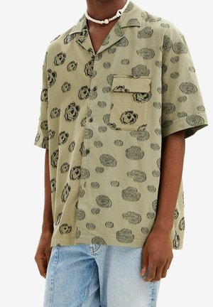 Homme portant une chemise ample à manches courtes de couleur vert olive avec des motifs floraux circulaires noirs et une poche frontale, associée à un jean en denim bleu clair.