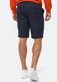 Helle marineblaue Shorts aus Baumwolle mit geradem Schnitt, ausgestattet mit zwei gesicherten Gesäßtaschen und einer weichen, glatten Textur. Kombiniert mit weißen Turnschuhen.